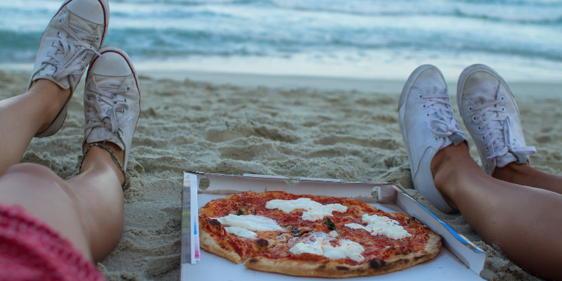 Two pairs of feet on either side of pizza box on the beach with ocean in the background.